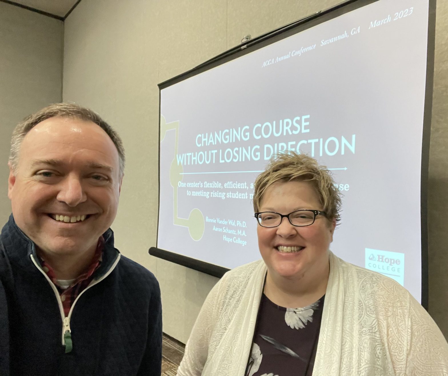 A close-up (selfie) of Aaron and Bonnie before their presentation. Aaron is on the left and Bonnie on the right with the screen in the background that says "Changing Course without Losing Direction" the title of their presentation.