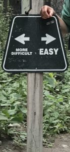 A closeup of a trapezoidal wooden trail sign.  It has two arrows in white.  The arrow pointing left says "more difficult" underneath, and the arrow pointing left says "easy" underneath.