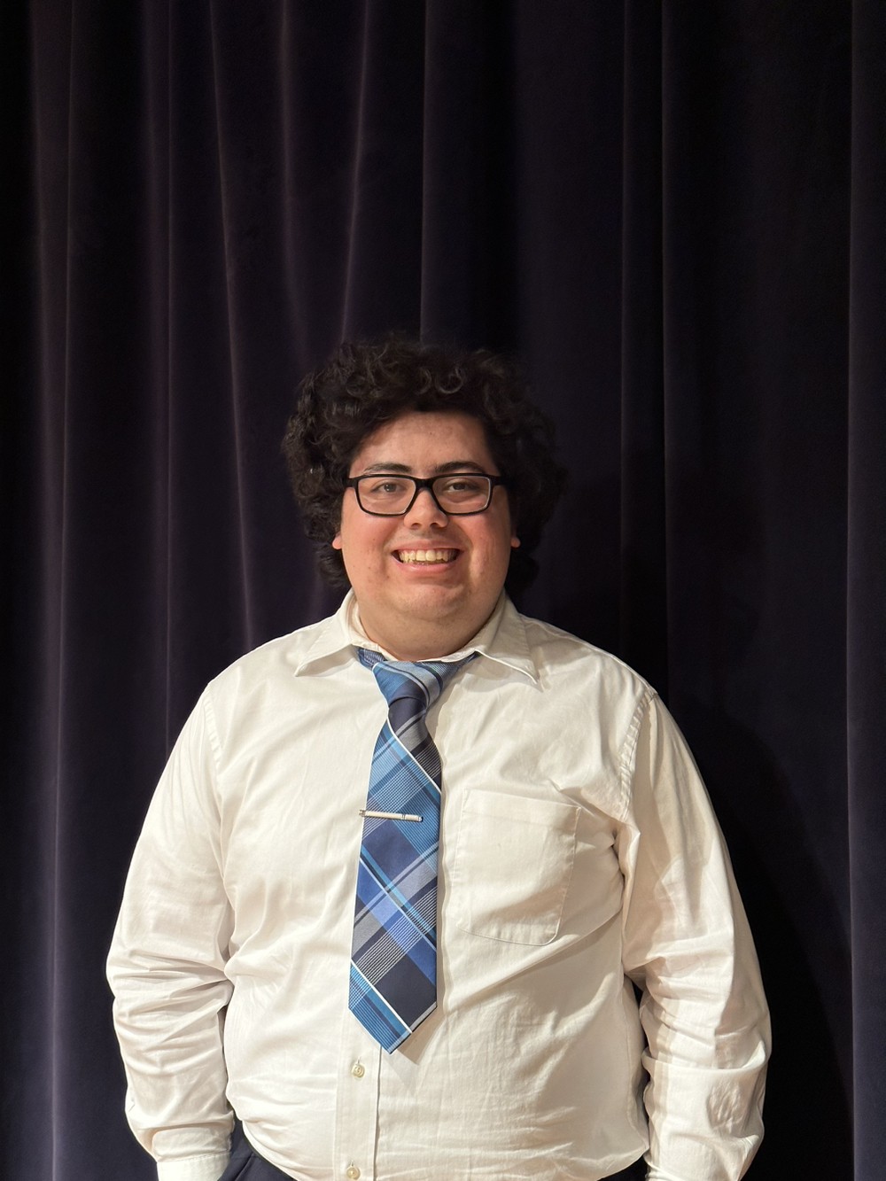 Latino student with glasses and dark curly hair in front of dark curtain in white shirt with blue patterned tie.