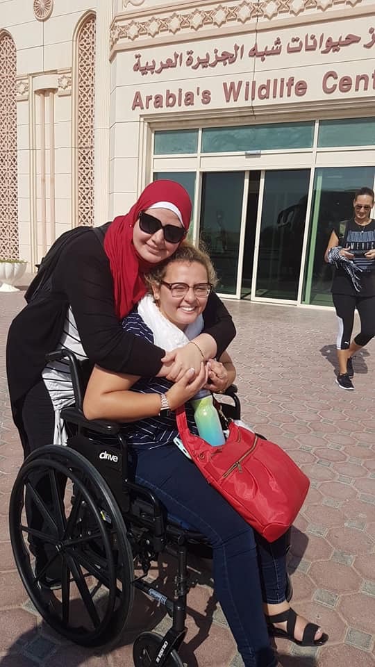 Safia is in her wheelchair in front of a building that reads "Arabia's Wildlife Center" in English with the Arabic directly above it. Hugging Safia from behind is a woman wearing sunglasses and a red hijab. They are both smiling