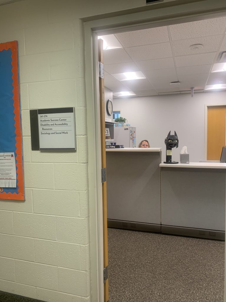A photo of the outside of disability services. The door is open and you can see Mrs. Eding's head peering over her desk. To the left, there reads a sign that says "261-264, Academic Success Center, Disability and Accessibility Resources, Sociology and Social Work".