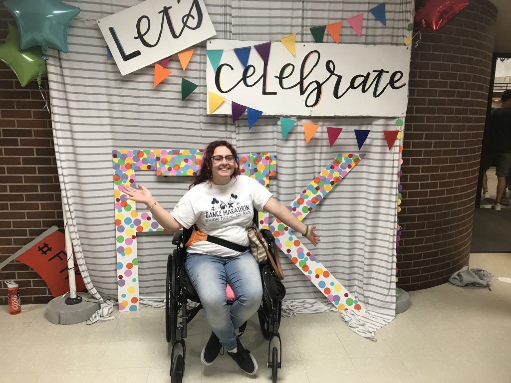 Safia is in her wheelchair wearing a Dance Marathon T-Shirt. She is sitting in front of a Dance Marathon background photo that is rainbow and says "Let's Celebrate" with a rainbow "FTK" leaning on the wall.