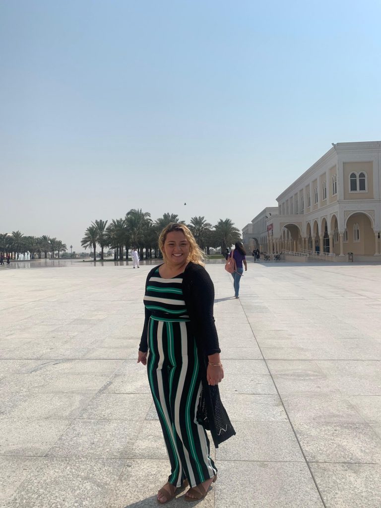 A picture of Safia standing outside the main quad at The American University of Sharjah. She is wearing a green, black, and white striped jumpsuit with a black cardigan. She is smiling, and the student center and date trees in the background.