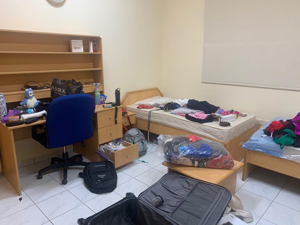 A picture of Safia's dorm room at the American University of Sharjah. There are clothes are various suitcase contents acorss the bed and on the desk in mid-unpacking. A suitcase lies on the floor mostly empty.