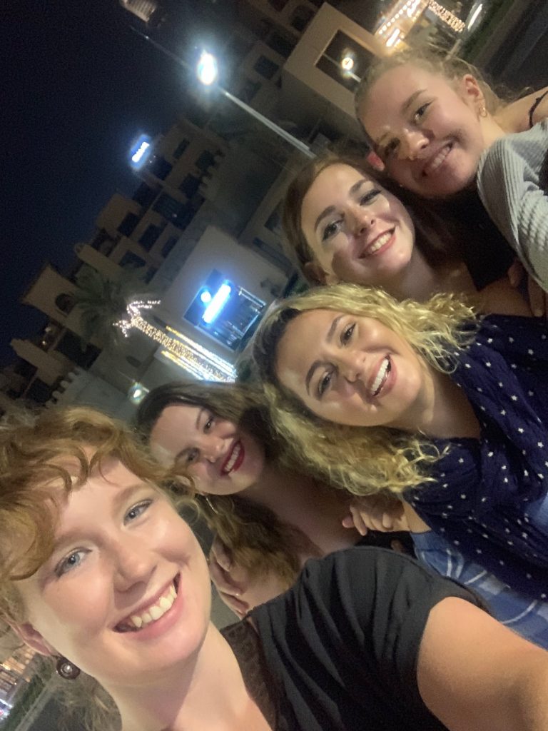 A selfie with five women (Alex, Tara, Taru, Alex, and Safia) is shown. They are standing outside some well-lit building in Dubai. They are all wearing makeup and are smiling.
