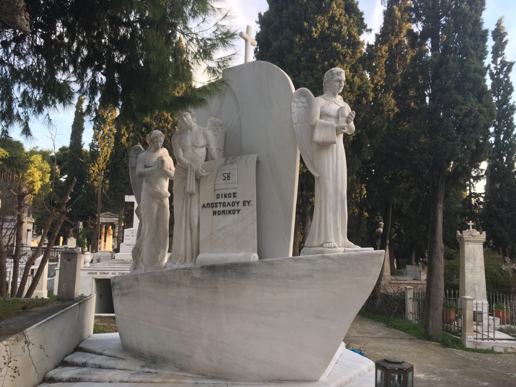 A carved marble boat with three figures. It is a headstone in a cemetery, and other graves can be seen in the background.