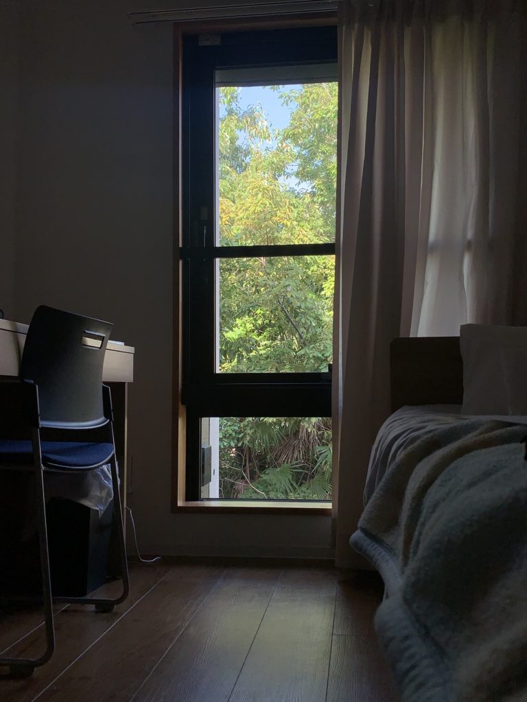 view of a window and curtain from inside the dorm room. on the left is a desk and chair and on the right is a bed.