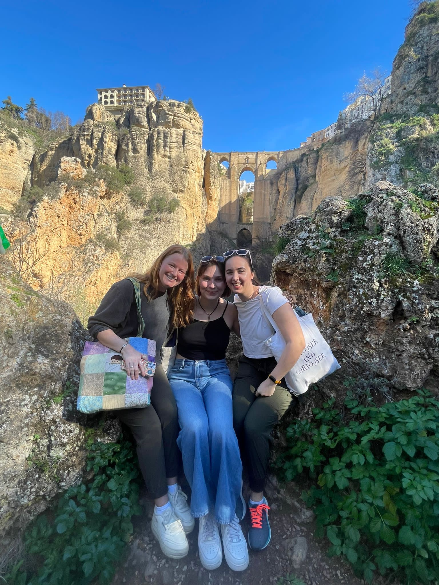 Three girls sitting on a rock with a large bridge behind them.