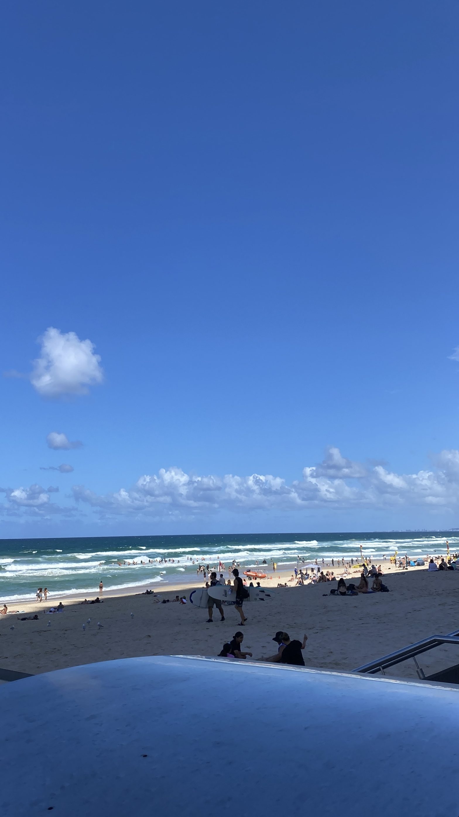 Beach scene with clear blue sky, fluffy clouds, and vibrant ocean waves. People relax on the sandy shore, some swimming, others surfing. Lively, sunny atmosphere.
