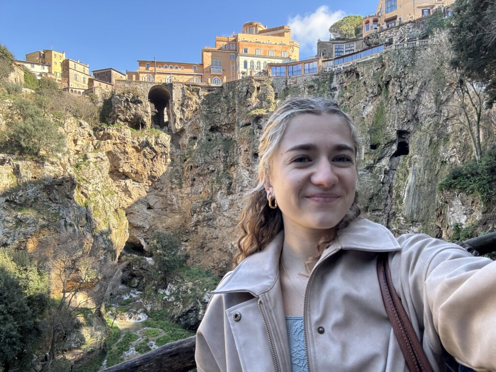A landscape-oriented selfie features Karley smiling in the foreground against a dramatic, mountainous backdrop in Tivoli. She is positioned on the right side of the frame, wearing a light beige or tan zip-up jacket over a soft blue top. Her wavy light-brown hair is partially pulled back, and she wears small gold hoop earrings. The background is a deep, rocky gorge with sheer stone cliffs. Perched precariously at the very top of these cliffs are several historic, sun-drenched buildings in shades of ochre and terracotta, overlooking the valley. A dark, arched tunnel or cave entrance is visible high up in the rock face. Below the buildings, a small, white-water stream cascades down through the greenery at the base of the ravine. The sky above is a crisp, bright blue, and the natural sunlight illuminates the rugged texture of the rocks and the greenery clinging to the slopes, creating a sense of ancient, scenic wonder.