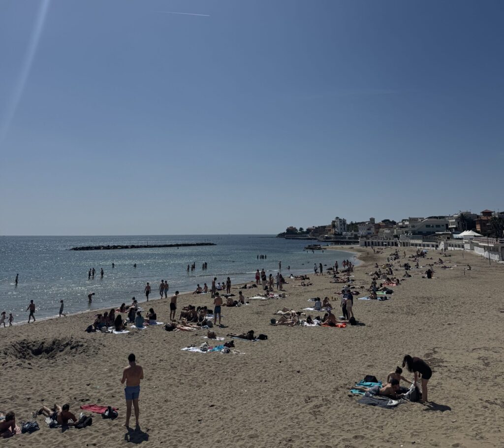 A high-angle shot captures a wide expanse of Santa Marinella beach under a brilliant, clear blue sky. The bright sun, positioned in the top-left corner, casts a long, vertical lens flare down the frame. The sandy beach curves gently toward the right, dotted with numerous people sunbathing on towels and walking along the shore. In the glistening blue water of the Tyrrhenian Sea, several swimmers can be seen enjoying the waves. A dark stone breakwater sits further out in the water to the left. Along the far right coastline, white buildings and beach clubs of the town line the shore. A faint white vapor trail from a plane cuts horizontally across the vast, empty sky, emphasizing the bright and open feel of a classic Italian spring day.