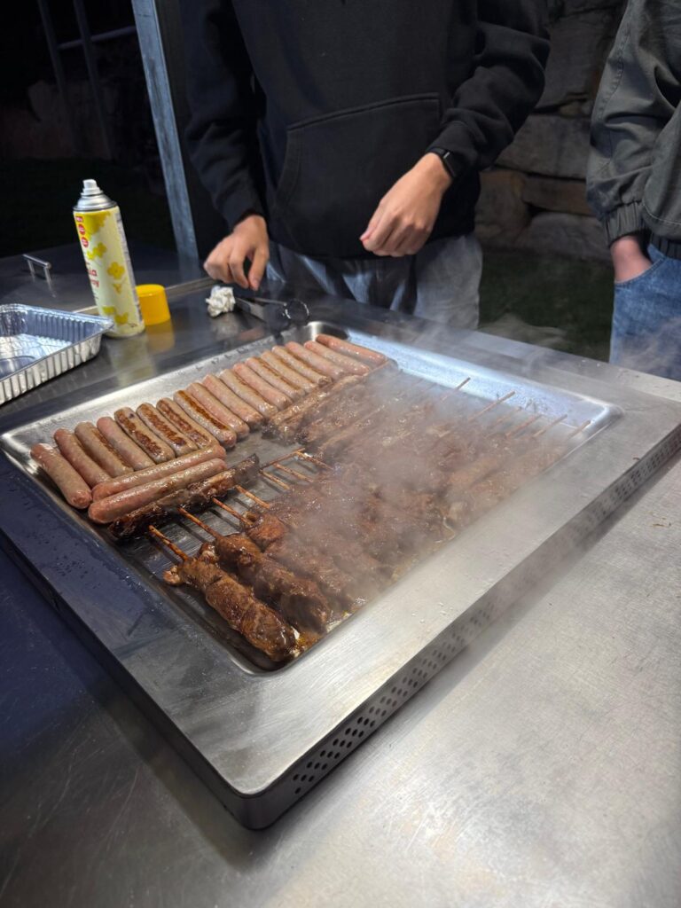 A group gathered around a flat-top grill outdoors, cooking rows of hot dogs and skewered meat, with steam rising and condiments placed nearby.