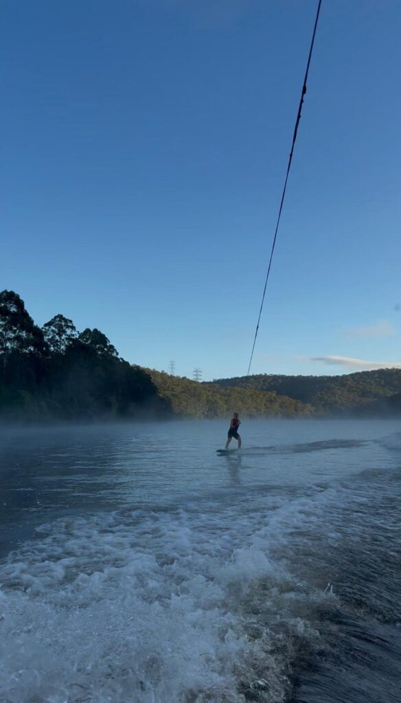 A girl wakeboarding on a river holding onto the rope connected to the boat with a background of green hills and lots of trees.