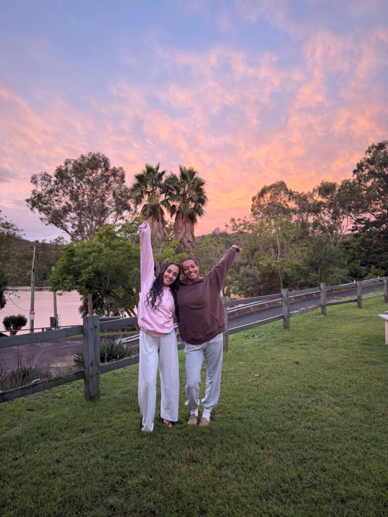 Two girls standing next to each other side hugging with one arm raised in front of a blue, orange, and pink sunset. One girl is wearing a pink sweatshirt and grey sweatpants, and the other girl is wearing a brown sweatshirt and grey sweatpants. They are standing on a grassy hill with a fence and some trees in the background.