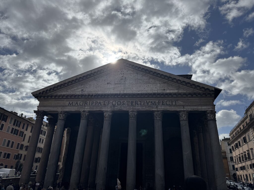 A wide-angle landscape shot captures the towering facade of the ancient Pantheon in Rome, Italy. The perspective looks up from the bustling piazza at the monumental portico, showcasing its massive Corinthian granite columns. The famous Latin inscription, “M·AGRIPPA·L·F·COS·TERTIVM·FECIT,” is clearly etched across the entablature beneath the triangular pediment. The sun is positioned directly behind the peak of the roof, creating a dramatic backlighting effect that casts the building's deep porch into heavy shadow while catching the edges of the stone in a bright glow. The sky above is filled with a dense pattern of white and grey clouds, with bright blue sky peeking through. Classic Roman apartments line the streets on the far left and right edges of the frame, grounding the ancient monument in the modern city.