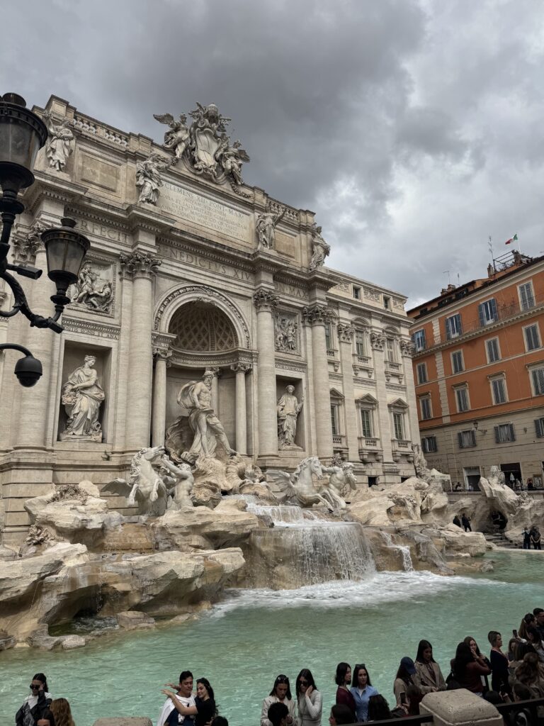 A vertical, medium-wide shot captures the iconic Trevi Fountain in Rome, Italy, under a heavy, overcast grey sky. The grand Baroque masterpiece dominates the left and center of the frame, showcasing its intricate white travertine marble facade. In the central niche, a powerful statue of Oceanus stands majestically atop a chariot shaped like a sea shell, pulled by taming seahorses and Tritons. Water cascades dynamically over the artificial rock formations at the base, creating a rushing waterfall that empties into the large, shimmering turquoise pool below. To the right of the monument, a classic five-story Roman apartment building stands in a warm terracotta-orange hue, its dark window shutters providing a colorful contrast to the pale marble of the fountain. In the lower foreground, a crowd of tourists can be seen gathering along the stone barrier at the edge of the pool, snapping photos and taking in the historic view.
