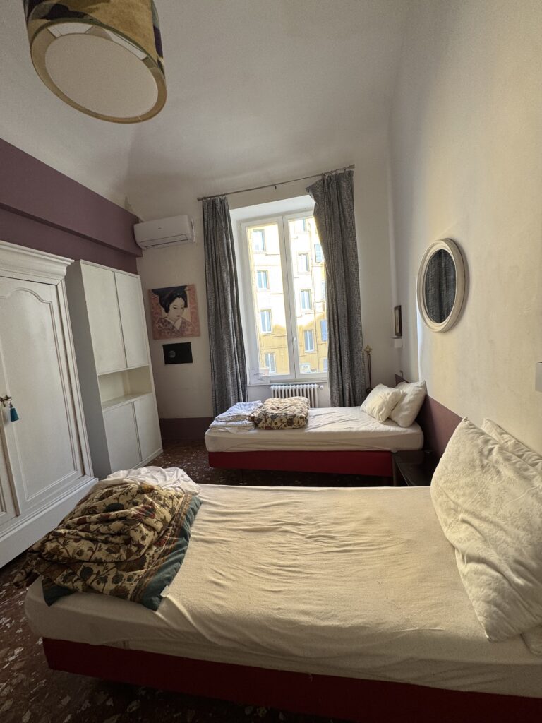 A vertical, wide-angle interior shot captures a cozy and simple European hotel room or guest house apartment.

The room is furnished with two single beds with red bases, positioned on opposite walls. The bed in the foreground has a rumpled white sheet and a folded, patterned tapestry-style quilt resting at the foot, while the second bed sits further back under a tall window.

The walls are predominantly painted off-white, accented by a deep plum-purple stripe running horizontally near the high ceiling and along the lower baseboards. To the left stands a large, white vintage wardrobe closet next to a simpler modern white storage unit.

In the center background, natural light streams in through a large double window draped with long, patterned grey curtains, offering a glimpse of a sunlit yellow building facade across the street. Decorative touches include a round mirror in a thick white frame mounted on the right wall, a small canvas print of a geisha, and a drum-style patterned lampshade hanging from the ceiling in the upper left foreground. The floor features dark, polished terrazzo tile, tying together the room's charming, historic feel.