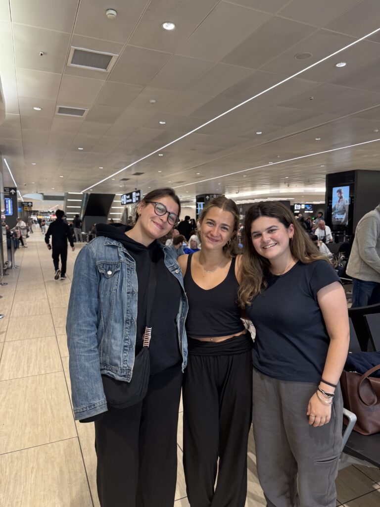 A vertical group portrait captures three smiling young women standing together inside a bright, modern airport terminal of the Rome Fiumicino Airport.

On the left, a young woman wearing round glasses, a black hoodie under a denim jacket, and black trousers stands with a black crossbody bag slung over her shoulder. In the center, a young woman with wavy light brown hair pulled back smiles warmly, dressed casually in a black tank top and flowing black pants. On the right, a young woman with long brunette hair smiles toward the camera, wearing a classic black t-shirt and grey sweatpants.

The background shows the sleek, wide expanse of the airport concourse, featuring polished light-tan flooring and long, recessed LED strip lighting stretching across the ceiling. In the distance, other passengers walk down the hall or wait near seating areas, and an airport digital display screen is visible on a pillar to the right, marking the winding down of an incredible European adventure.