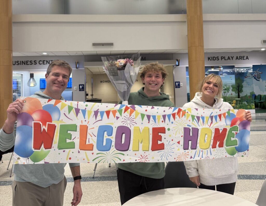 A shot captures the ultimate homecoming moment inside the bright, modern arrivals terminal at Gerald R. Ford International Airport (GRR) in Grand Rapids. Standing behind a white circular table, a three-person welcome committee beams at the camera, proudly holding up a large, colorful "WELCOME HOME" banner adorned with festive balloons, confetti, and streamers. On the left, a man in a grey hoodie and khaki pants holds one end of the banner with a wide smile, while in the center, a young man in a green hoodie holds the middle of the sign while balancing a beautiful bouquet of fresh flowers. On the right, a young woman with blonde bangs tilts her head happily, wearing a cozy oversized white sweatshirt and holding the other end of the banner. The clean, open architecture of the terminal forms the background, featuring tall wooden-paneled pillars, a slatted wood ceiling, and signs for the business center and kids' play area visible in the distance, completing a heartwarming end to a long journey.