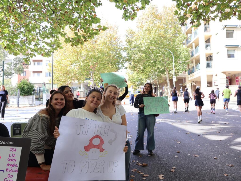 A group of friends standing along a tree-lined Sydney street holding colorful handmade signs and cheering during the half marathon race.