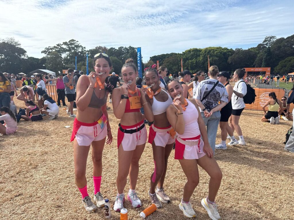 Four friends in coordinated pink running outfits smiling and showing off their medals after finishing the half marathon among a crowd of runners.