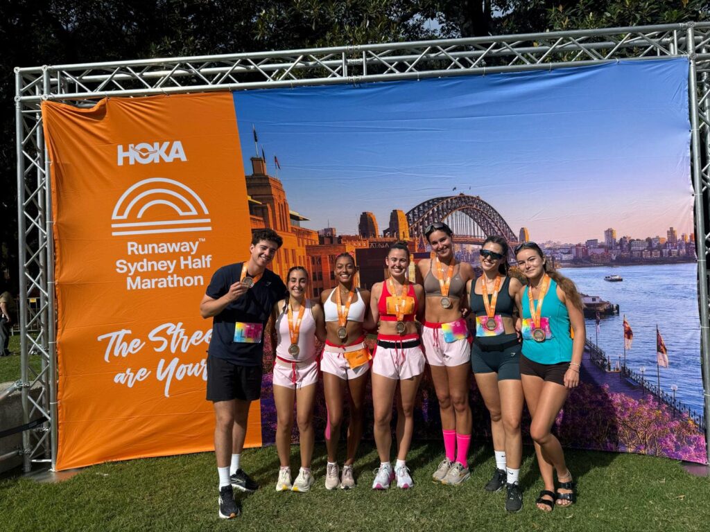 A group of runners posing together with medals after completing the HOKA Raceway Half Marathon, with the Sydney Harbour Bridge also on the banner in the background.
