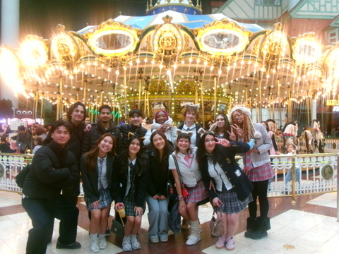 A group of people poses in front of a brightly lit caroseul. Several are smiling, or making peace signs. The illuminated carousel in the background creates a festive and cheerful atmosphere.