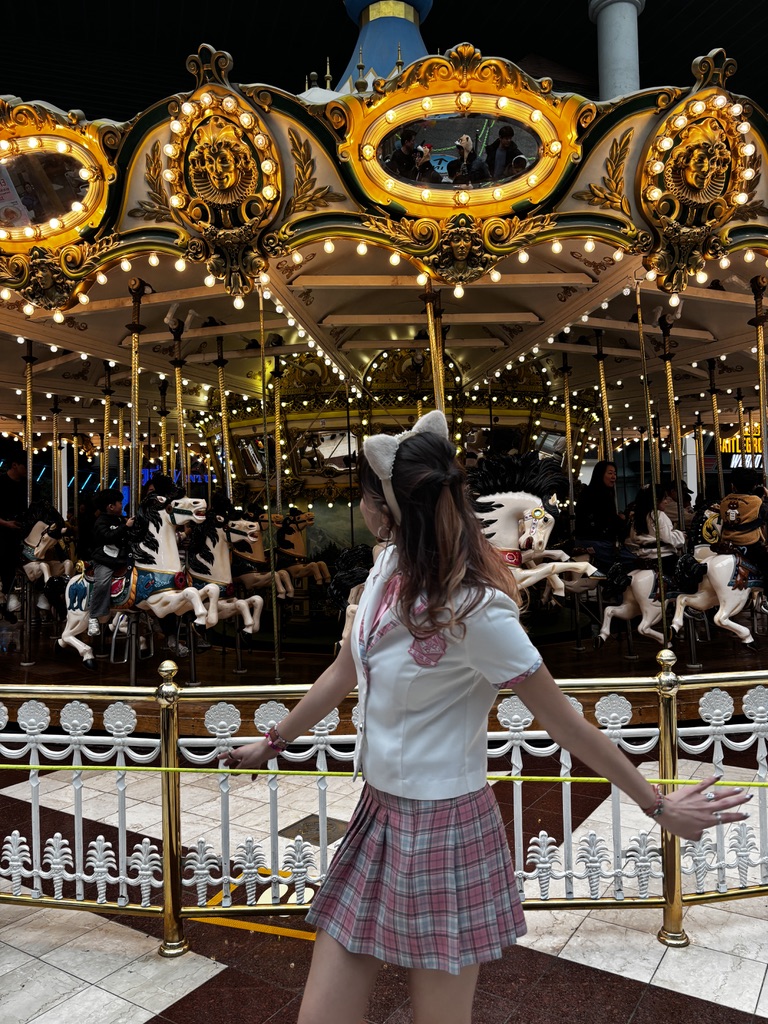A person standing in front of a brightly lit carousel, wearing a white top, pink plaid skirt, and a cat-ear headband.