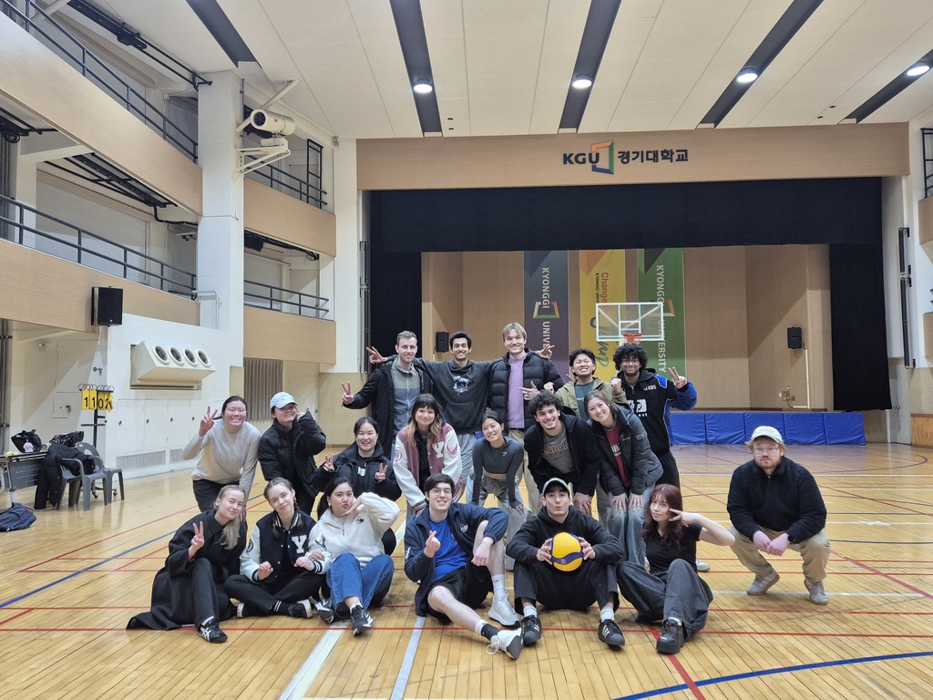 A group of students posing together on an indoor gymnasium court, with a volleyball, under a banner reading "KGU" and a basketball hoop in the background.