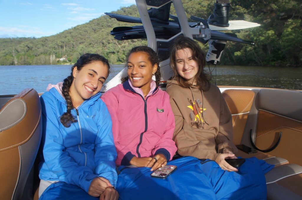 Three friends smiling while sitting together on a boat, with green hills and calm water in the background.