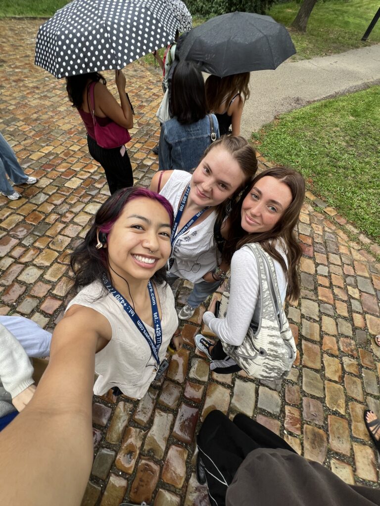 3 smiling young women pose for a selfie on a brick path, with two others holding umbrellas behind them. The scene feels cheerful and rainy.