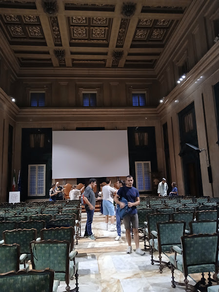 Photo of a large room with very high ceilings and classical, decorative moldings on the ceiling and high on the walls. Rows of upholstered chairs in wood frames are lined up, and the photo is taken from behind the chairs. The chairs face a small stage with a very large screen hung above it. Nine to ten people are milling around, and some seem to be walking out of the room.