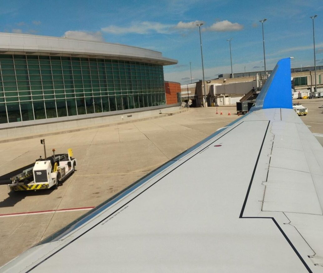 Airplane wing with a bright blue wingtip on an airport tarmac. The photo is taken from a window seat near the wing, showing the long white wing stretching toward the distance, with flap panels visible along its trailing edge. Below the wing is a concrete apron with a red guideline and some darker stained areas. Near the center-bottom of the image there is a small white and yellow ground vehicle positioned close to the wing, with a tow bar extended toward the aircraft. To the left side of the scene is an airport terminal building with a curved roof and a large glass facade made up of green-tinted window panels framed by white structural elements. Further left, tall light poles rise above the terminal area against a mostly clear blue sky with a few thin clouds. Description provided by Be My Eyes AI.