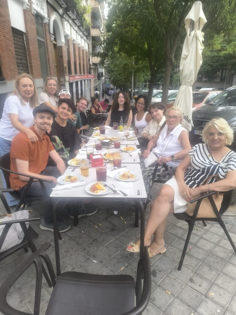 A group of eleven people sitting around a rectangular outdoor café table on a city sidewalk. They are arranged on three sides of the table, all facing toward the camera and smiling, giving a relaxed, cheerful feeling. On the left side, two older women stand just behind the seated people, leaning in to be part of the group. In front of them, a young man in an orange shirt and flat cap sits at the near left corner, turned slightly toward the center. Along the far side of the table, several younger people sit close together, with casual clothing and varied hairstyles, including curly and straight hair; some rest their arms on the table, and one person has an arm around another’s shoulder. On the right side of the table, two older women sit nearest the camera. The one closer to the center wears a white sleeveless top and light pants with glasses and a cross‑body bag. The woman at the near right corner wears a white skirt and a black‑and‑white striped top, with a tan handbag hanging from the chair and legs crossed. The table is covered with white placemats and has small plates of food such as what appears to be pieces of tortilla or egg dishes with toppings, plus drinks including light beer‑colored and red beverages in glasses. There is a small red box or container in the center of the table, along with condiment bottles. The café chairs are black with armrests, and one empty chair is in the foreground. In the background, there are parked cars along the street, tall leafy trees providing shade, and a row of brick and stone buildings with shops at street level. An upright closed patio umbrella stands beside the group on the right. The atmosphere suggests a casual group meal or gathering at an outdoor restaurant in an urban neighborhood. Summary provided by Be My Eyes AI