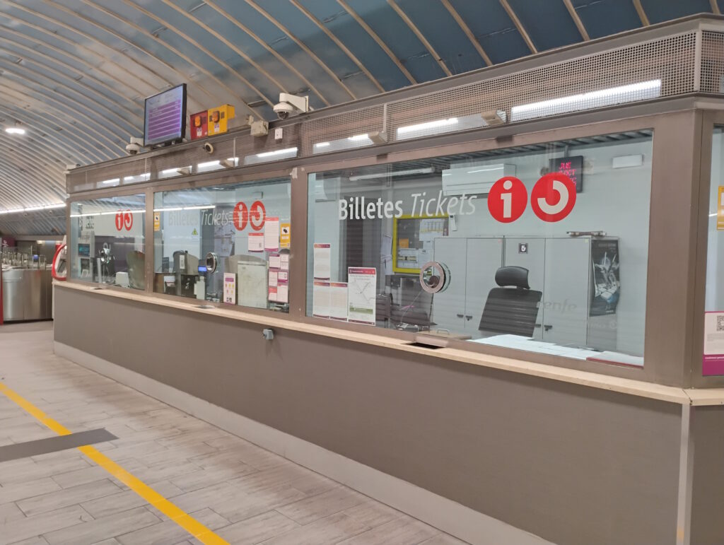 Ticket office windows in a train or metro station. A long counter runs from the foreground into the distance, with large glass windows separating the customer side from the staff area. On the glass, white text reads “Billetes Tickets” along with several large red circular symbols containing a lowercase “i” for information. Behind the glass, there are office chairs, cabinets, a computer monitor, and various posters and notices pinned up. Above the windows, electronic equipment and screens are mounted high on the wall, including a display showing information in pink and white. The ceiling has curved, ribbed panels in a light blue color, arching over the area. On the floor in front of the counter, light-colored tiles are marked with yellow lines guiding where people should walk or queue.