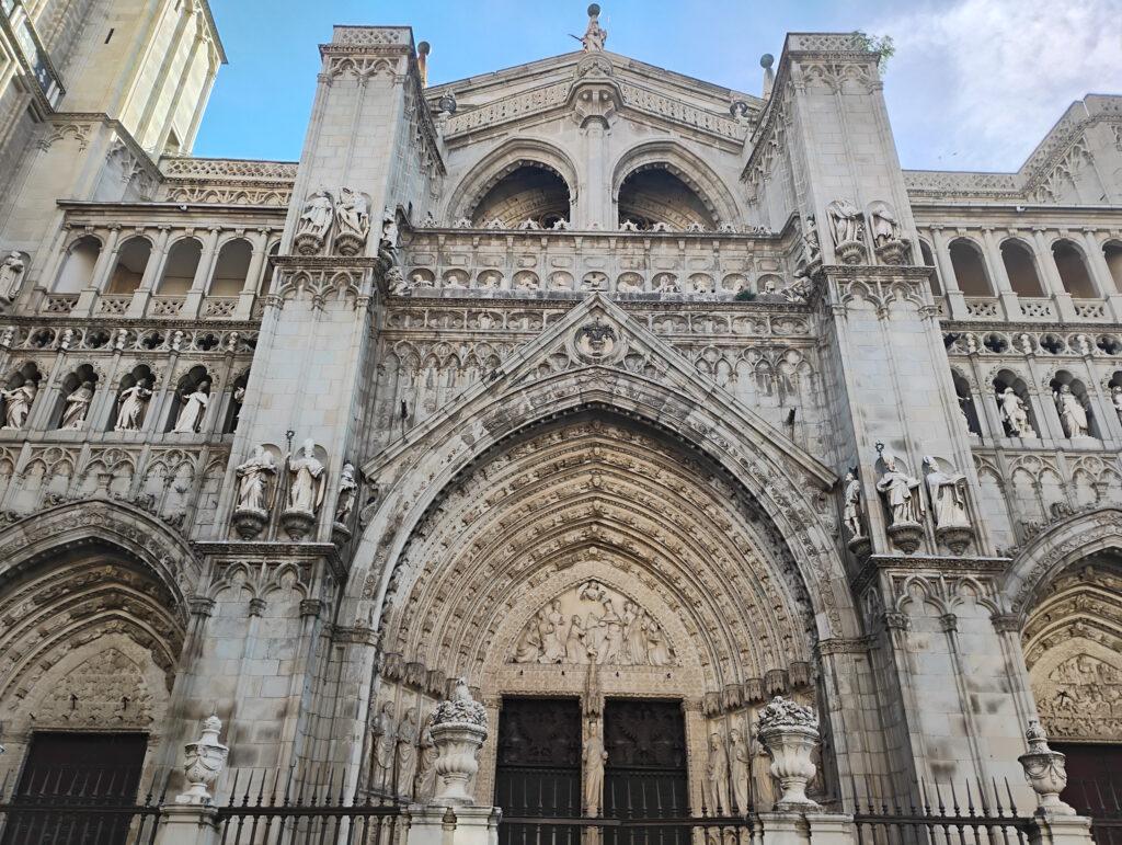 Gothic cathedral façade with a large central entrance and two side entrances, all covered in ornate stone carvings. The structure is made of pale stone and rises vertically with strong, tall pillars on the left and right sides of the main entrance. The central portal is framed by multiple pointed stone arches that recede inward, each arch ring densely filled with small sculpted figures and decorative patterns. Above the main doorway, in the tympanum area, a detailed relief scene shows many human figures arranged in rows; smaller carved scenes appear in the arches over the two side doors as well. Flanking the main entrance, tall statues of standing figures in long robes occupy niches in the columns, and similar statues run in a continuous row across the façade on an upper level. Higher up, another large pointed arch frames an open gallery with more sculpted figures inside, and additional layers of intricate tracery and small carvings fill almost every surface. The very top of the façade forms a triangular gable with more stone detailing and a round medallion-like carving at the center. Dark wooden doors fill the three portals at ground level, partially hidden behind a black metal fence with vertical spikes. The sky above is clear with a light blue tone, giving strong contrast to the light-colored stone of the cathedral. Summary provided by Be My Eyes AI.