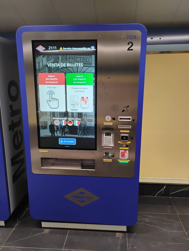 This is a blue Metro ticket machine with a large touch screen in the center. Above the screen, from left to right: Card slot for bank cards, Cash note slot, Contactless card area, Ticket/card slot with a green light, Keypad with a small display, Ticket/receipt output slot. On the screen you can choose ticket options and languages, and there is an accessible-use button with a wheelchair symbol. Summary provided by Be My Eyes AI.