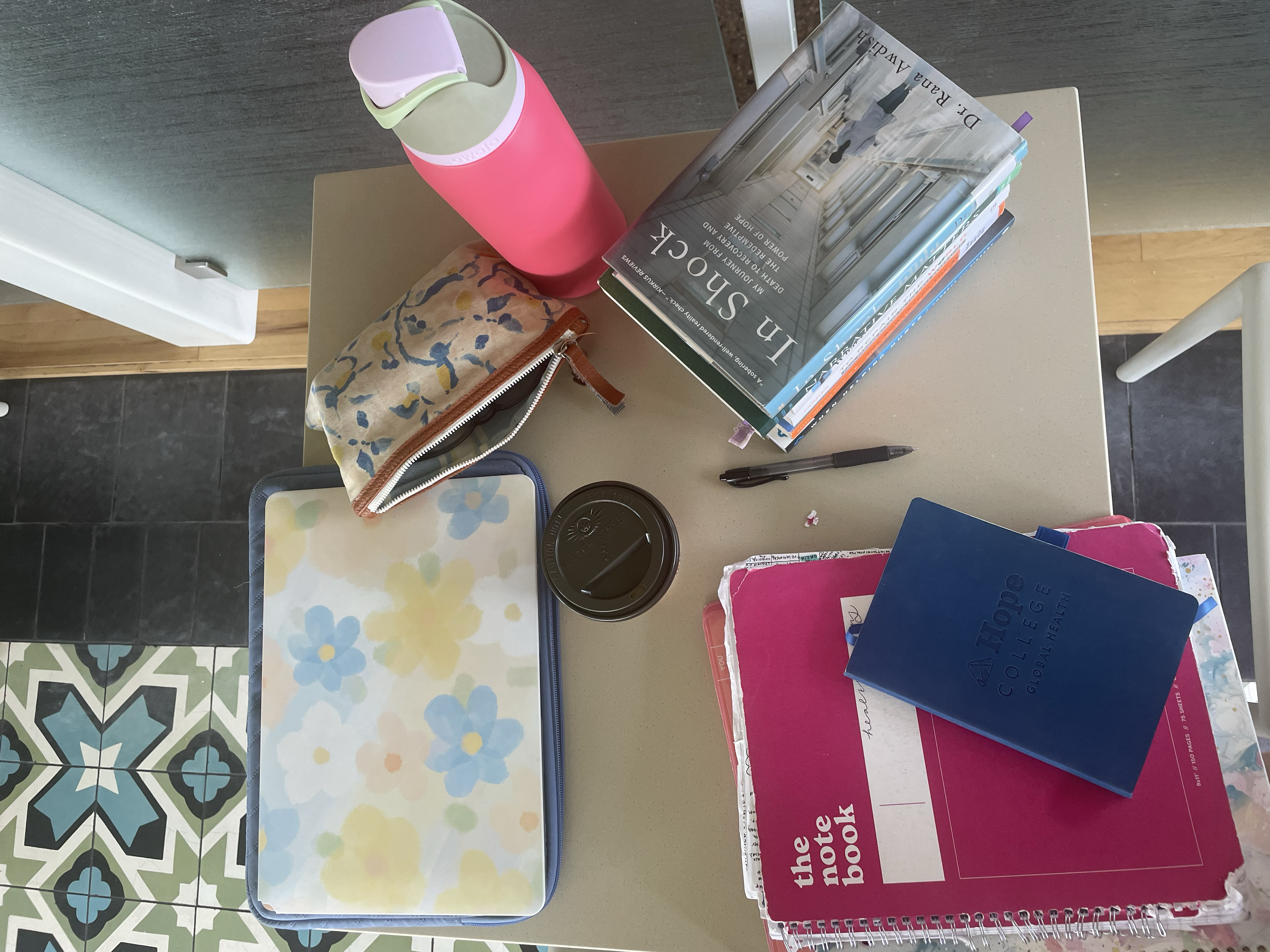 Overhead view of a dskw with a stack of books, notebook, pink water bottle, ink pen and pencil pouch layin on it.